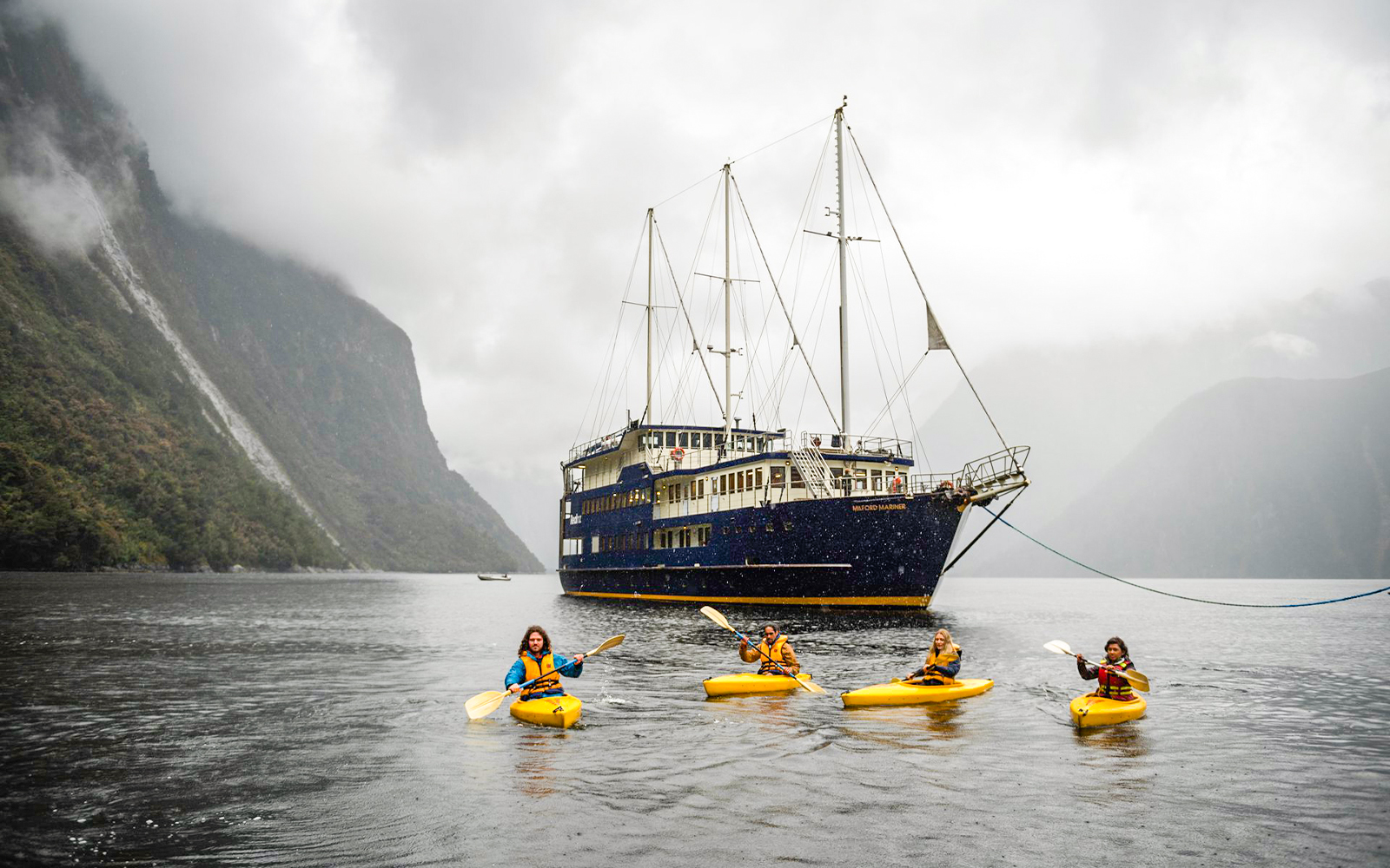 Kayakers paddle near a cruise ship in Milford Sound, New Zealand, surrounded by misty mountains.