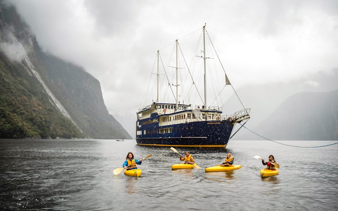 Kayakers paddle near a cruise ship in Milford Sound, New Zealand, surrounded by misty mountains.