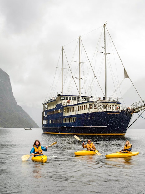 Kayakers paddle near a cruise ship in Milford Sound, New Zealand, surrounded by misty mountains.