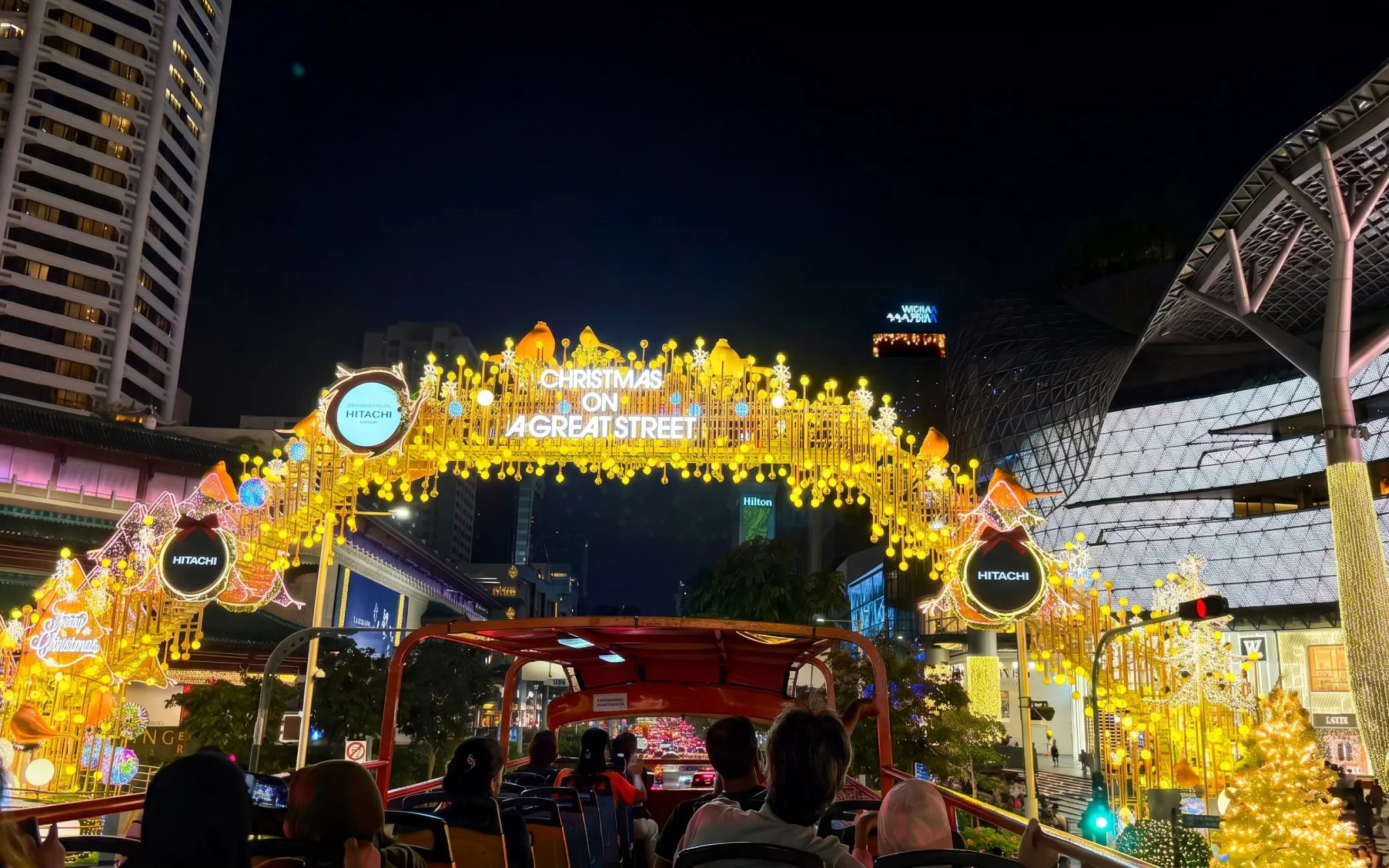 Tour bus passengers under illuminated "Christmas on a Great Street" arch in cityscape at night.