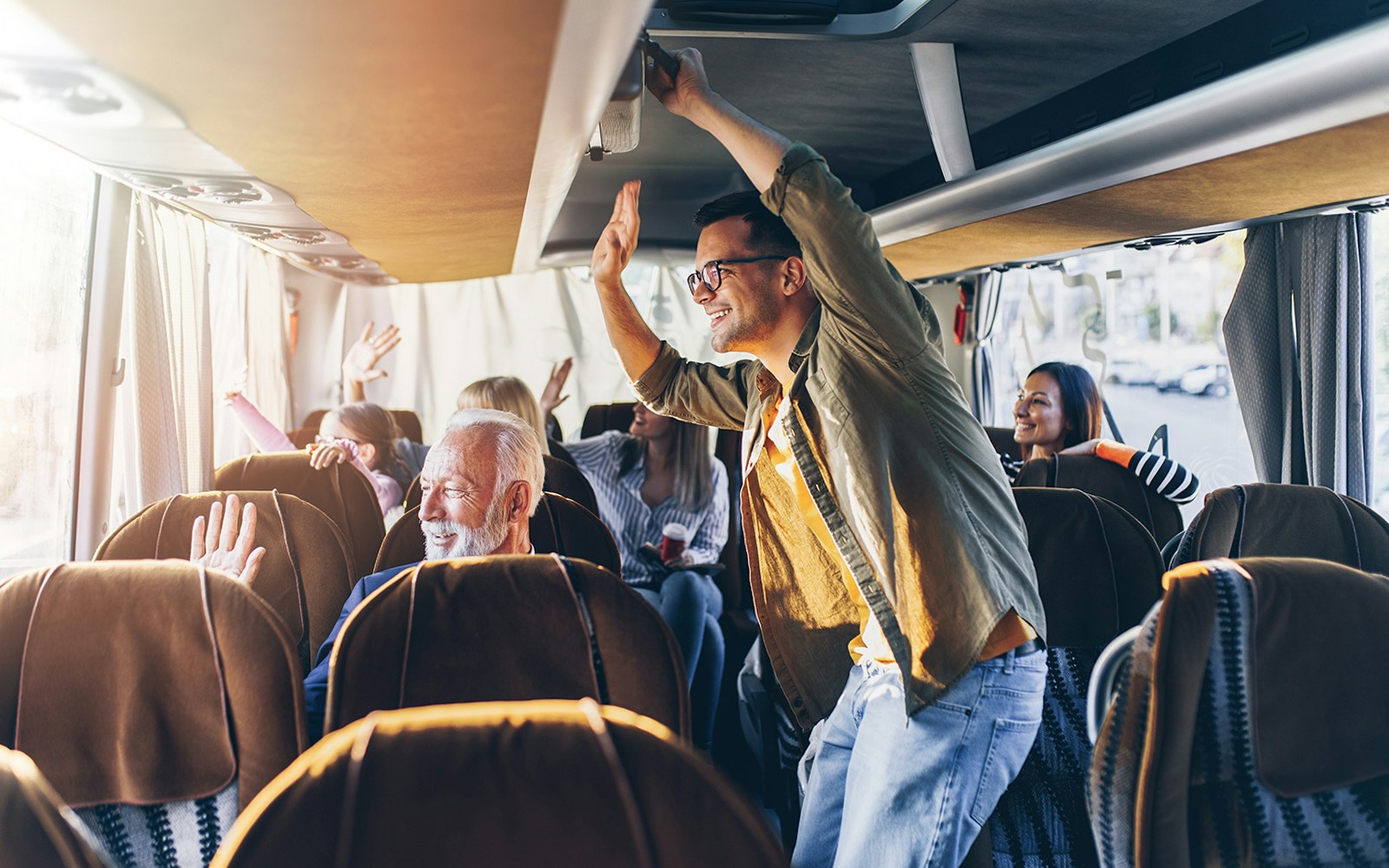 Passengers enjoying a bus ride, some standing and waving, with sunlight streaming through windows.