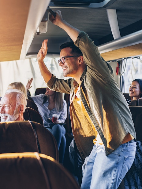 Passengers enjoying a bus ride, some standing and waving, with sunlight streaming through windows.