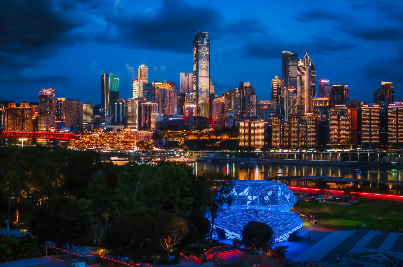 Chongqing skyline at night with illuminated skyscrapers and river view, seen from a park.