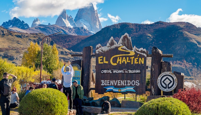 Tourists at El Chaltén welcome sign with mountains in the background, Argentina.