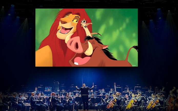 Orchestra performing with "Lion King" characters Simba, Timon, and Pumbaa on screen.