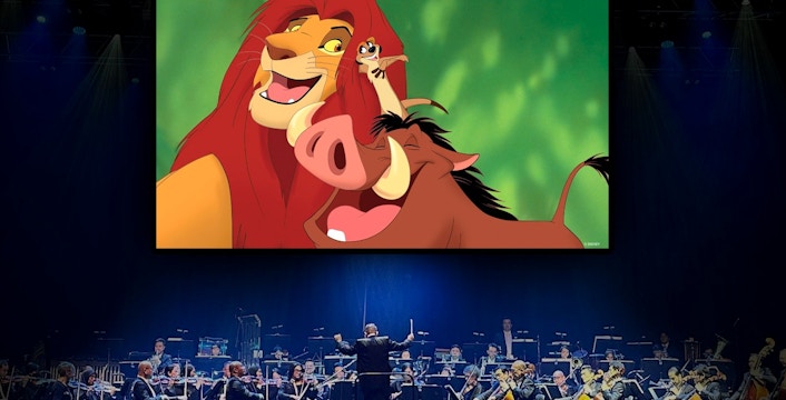 Orchestra performing with "Lion King" characters Simba, Timon, and Pumbaa on screen.
