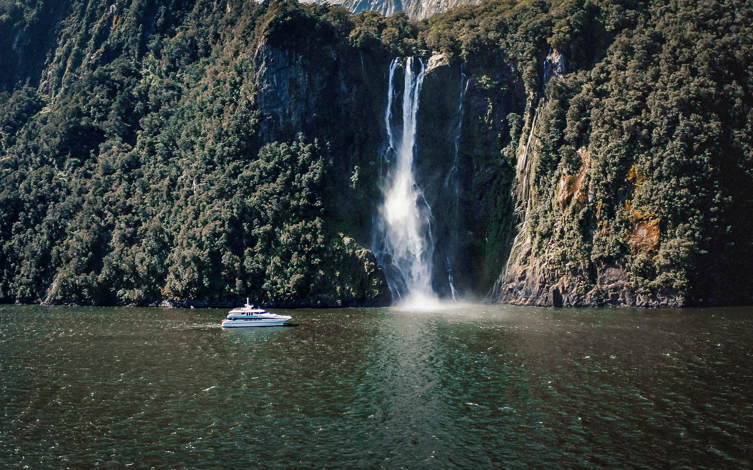 Cruise boat near waterfall in Milford Sound, New Zealand, surrounded by lush cliffs.