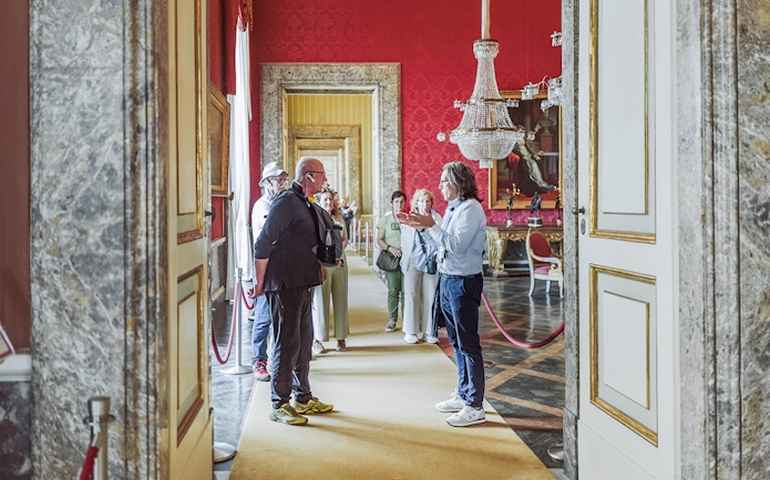 Tour group with guide inside Royal Palace of Caserta, ornate red room with chandelier.