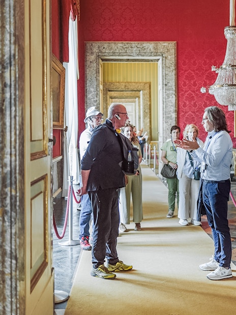 Tour group with guide inside Royal Palace of Caserta, ornate red room with chandelier.