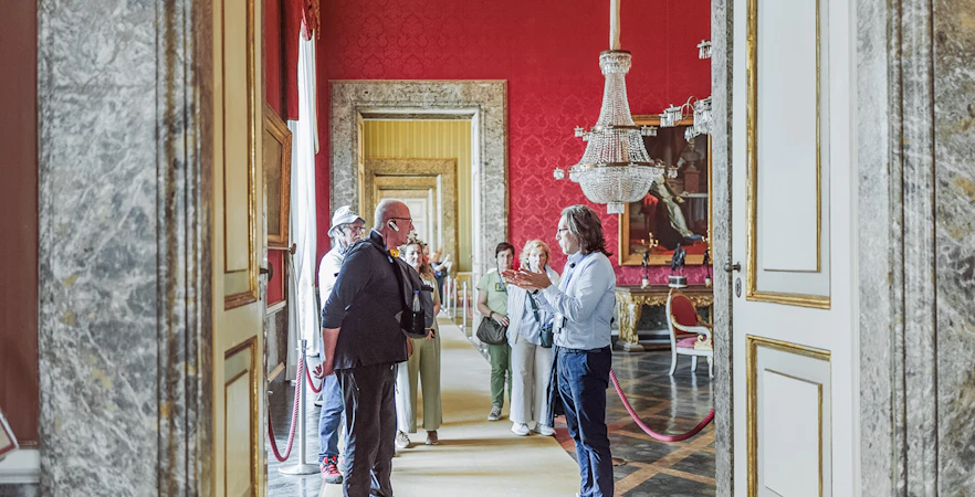Tour group with guide inside Royal Palace of Caserta, ornate red room with chandelier.