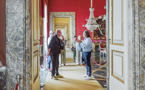 Tour group with guide inside Royal Palace of Caserta, ornate red room with chandelier.