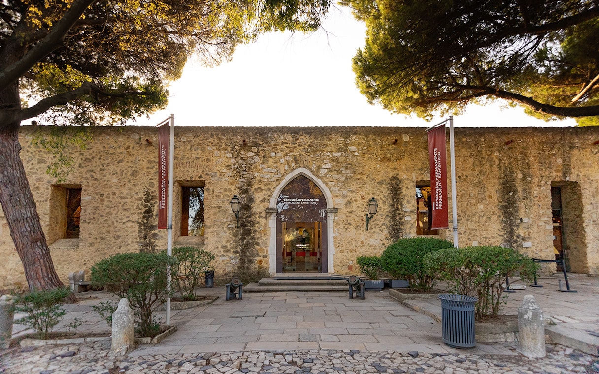 Entrance to Permanent Exhibitions at St George's Castle, Lisbon, with stone facade and flags.