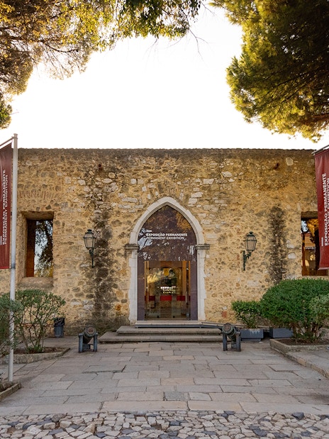 Entrance to Permanent Exhibitions at St George's Castle, Lisbon, with stone facade and flags.