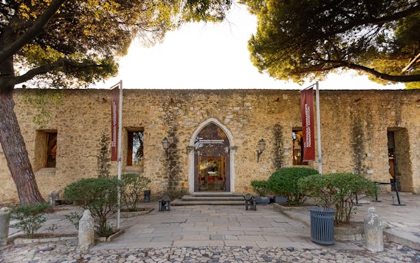 Entrance to Permanent Exhibitions at St George's Castle, Lisbon, with stone facade and flags.