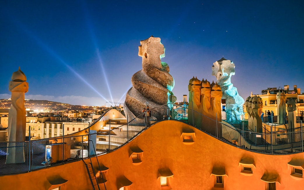 Casa Mila rooftop at night with illuminated chimneys and Barcelona skyline.