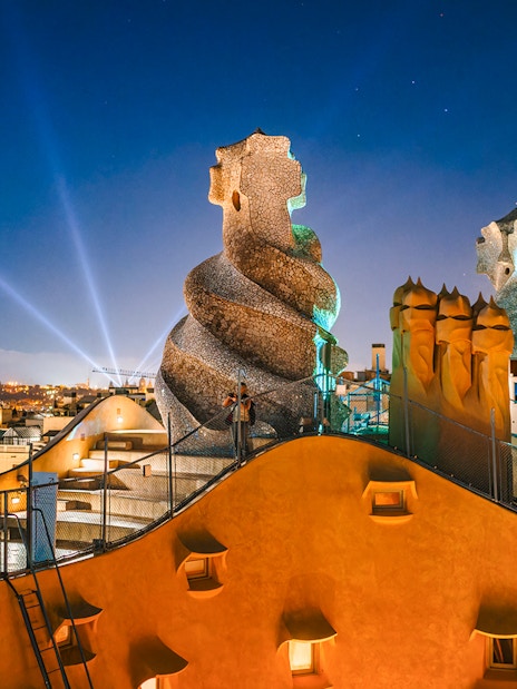 Casa Mila rooftop at night with illuminated chimneys and Barcelona skyline.