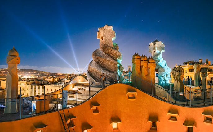 Casa Mila rooftop at night with illuminated chimneys and Barcelona skyline.