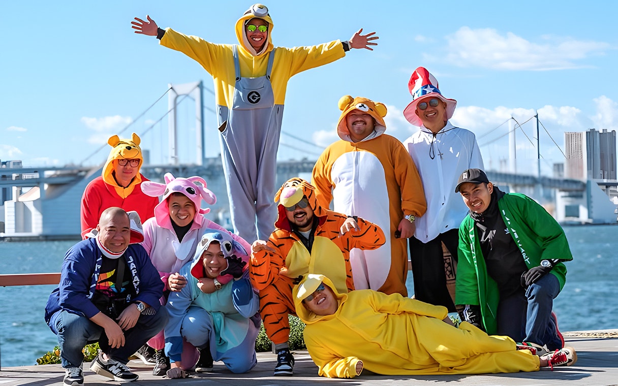Group in colorful costumes posing by Osaka waterfront with bridge in background.