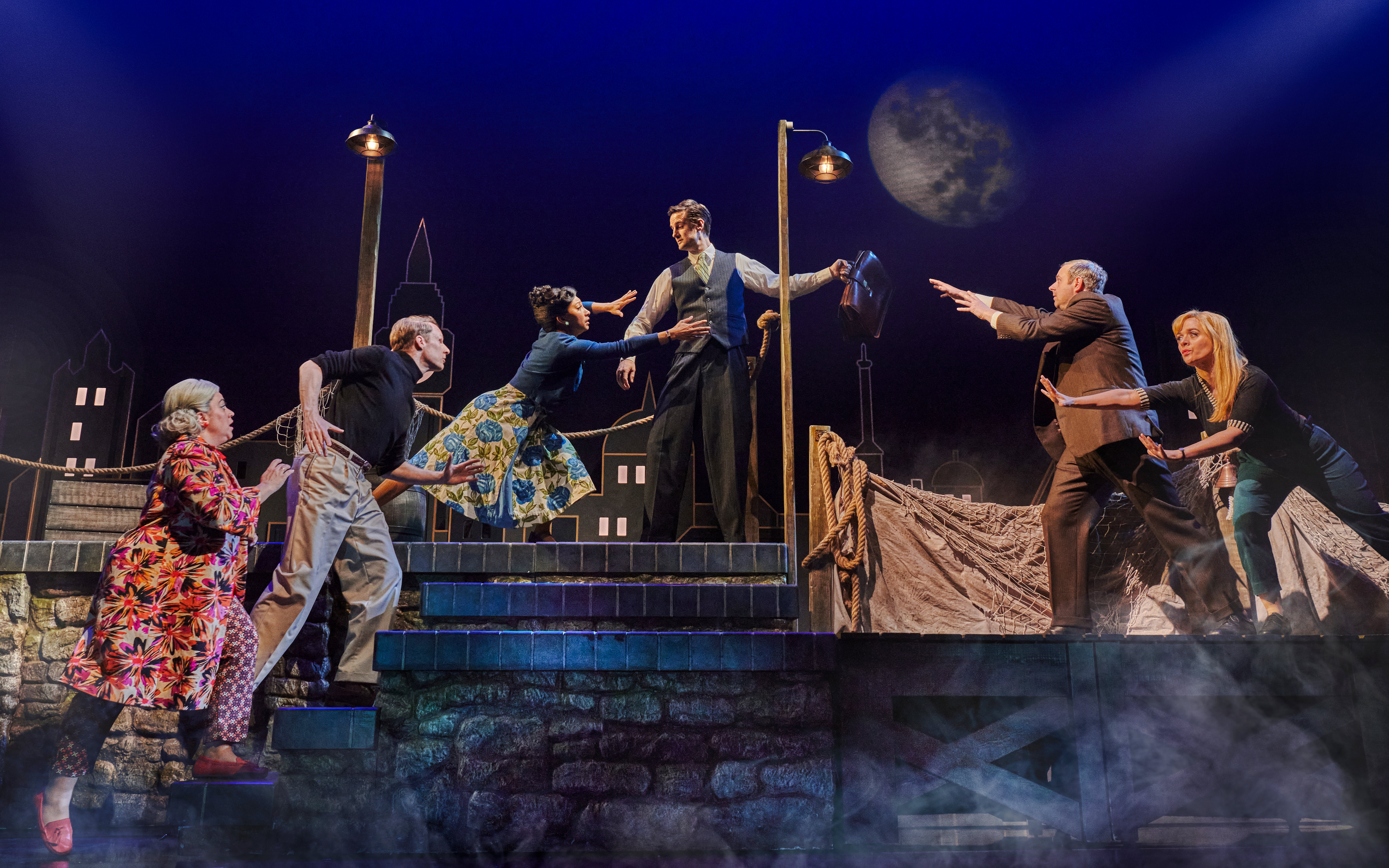 Actors performing a dramatic scene in "The Comedy About Spies" with a cityscape backdrop.