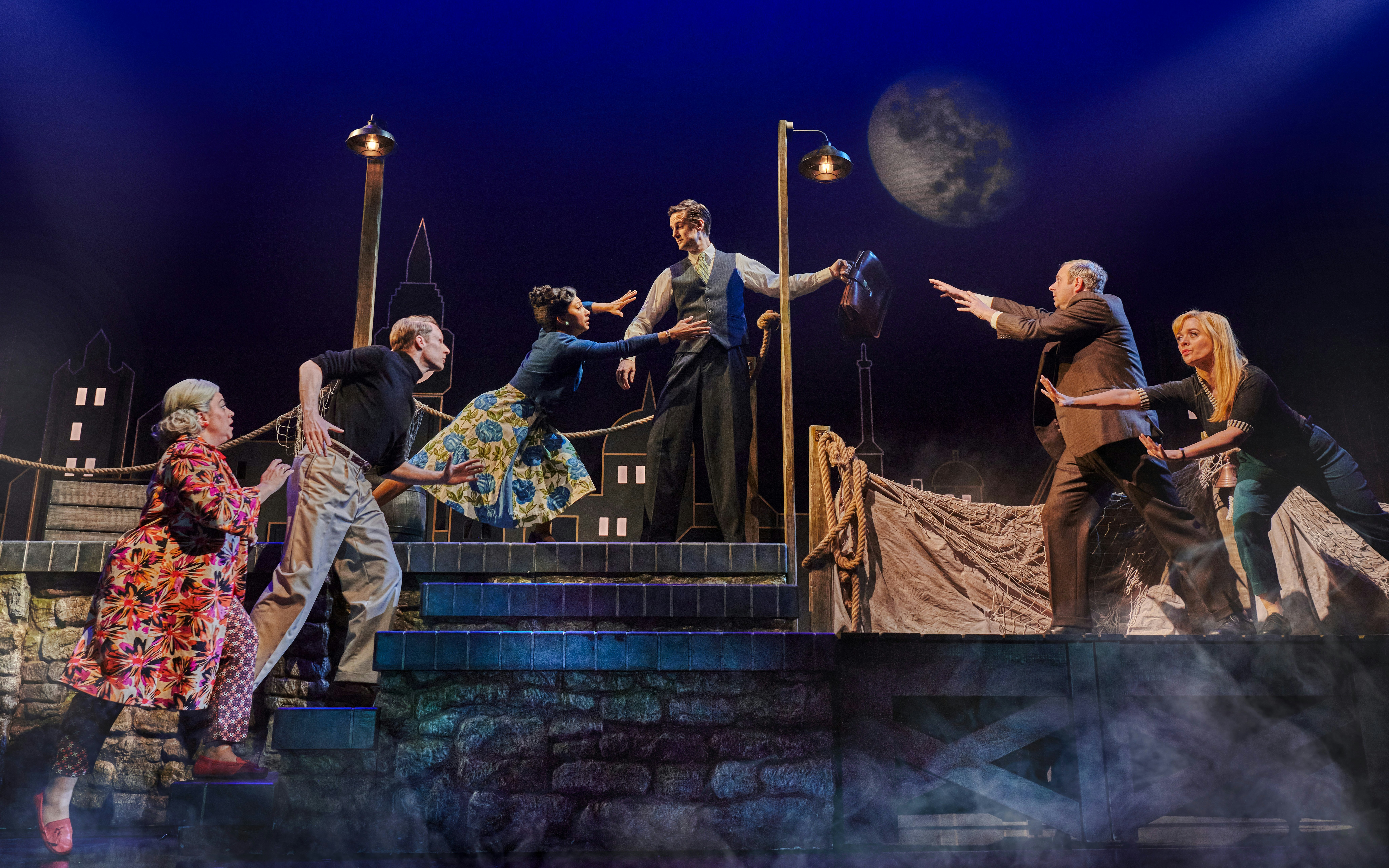 Actors performing a dramatic scene in "The Comedy About Spies" with a cityscape backdrop.