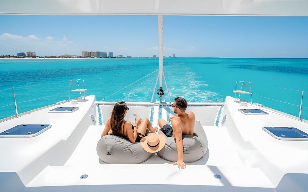 Tourists relaxing with drinks on a catamaran in Isla Mujeres, turquoise sea in background.