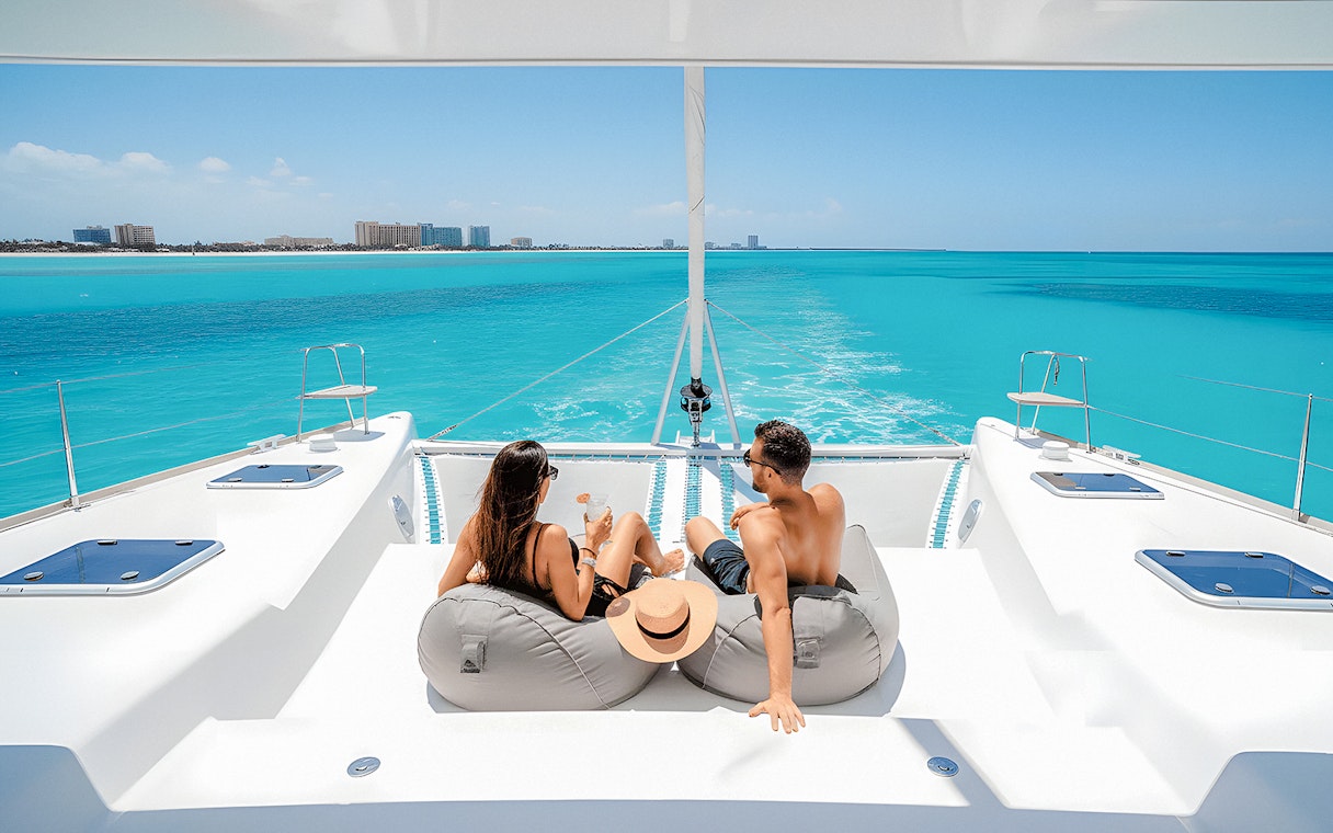 Tourists relaxing with drinks on a catamaran in Isla Mujeres, turquoise sea in background.