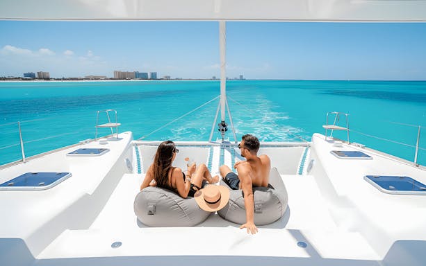 Tourists relaxing with drinks on a catamaran in Isla Mujeres, turquoise sea in background.
