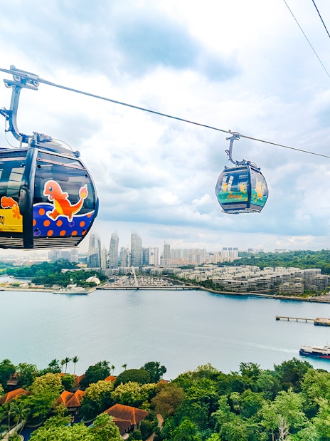 Cable car with Pokémon theme over Sentosa, Singapore, with city skyline and water view.