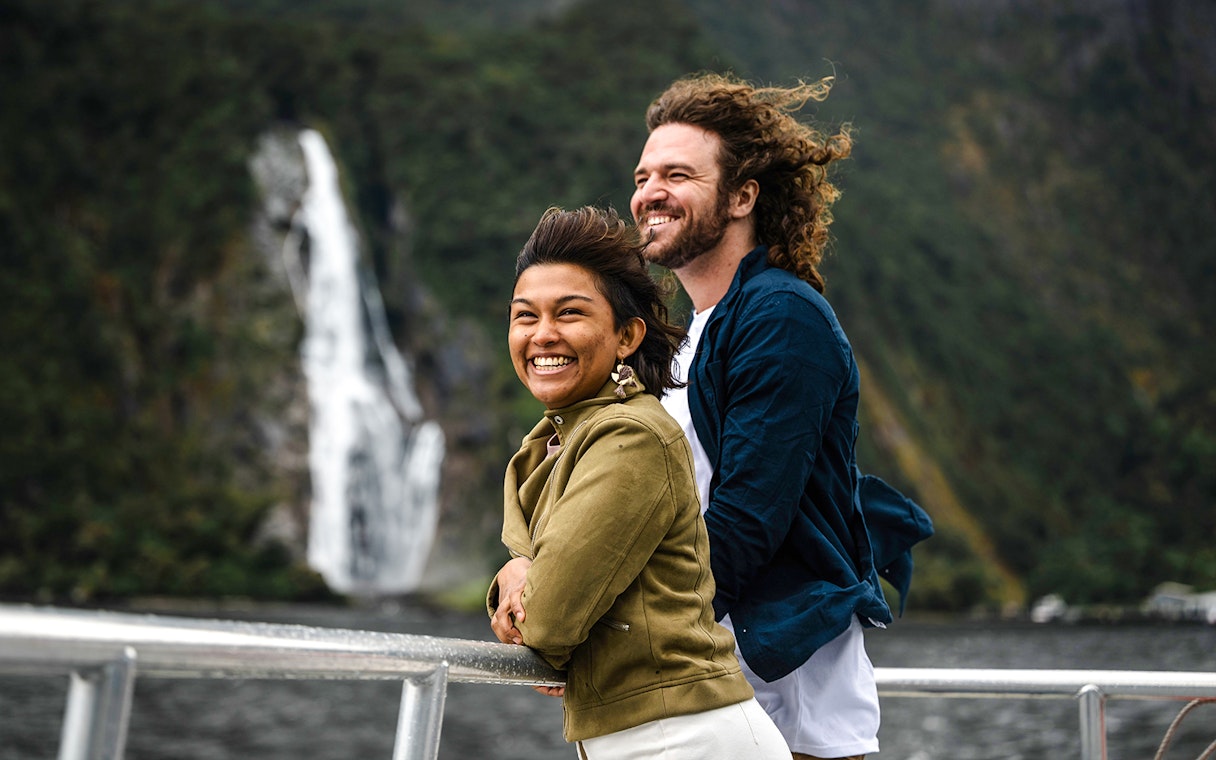 Couple enjoying Milford Sound cruise from viewing deck with waterfall in background.