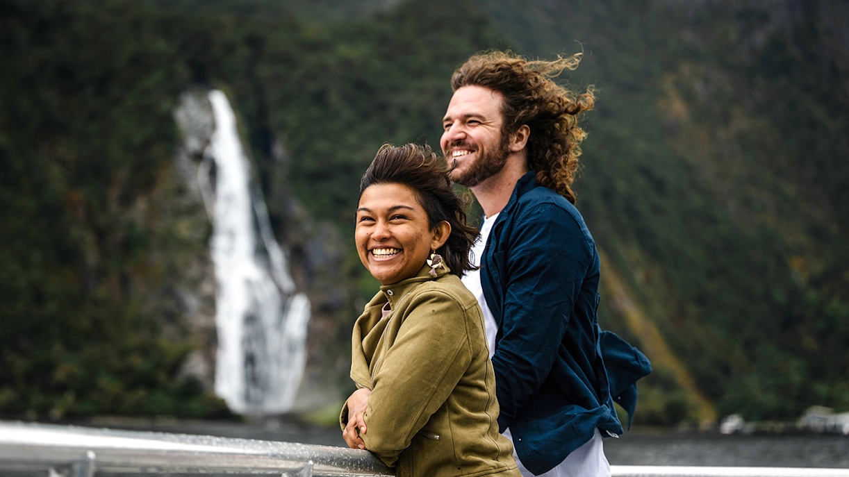 Couple enjoying Milford Sound cruise from viewing deck with waterfall in background.