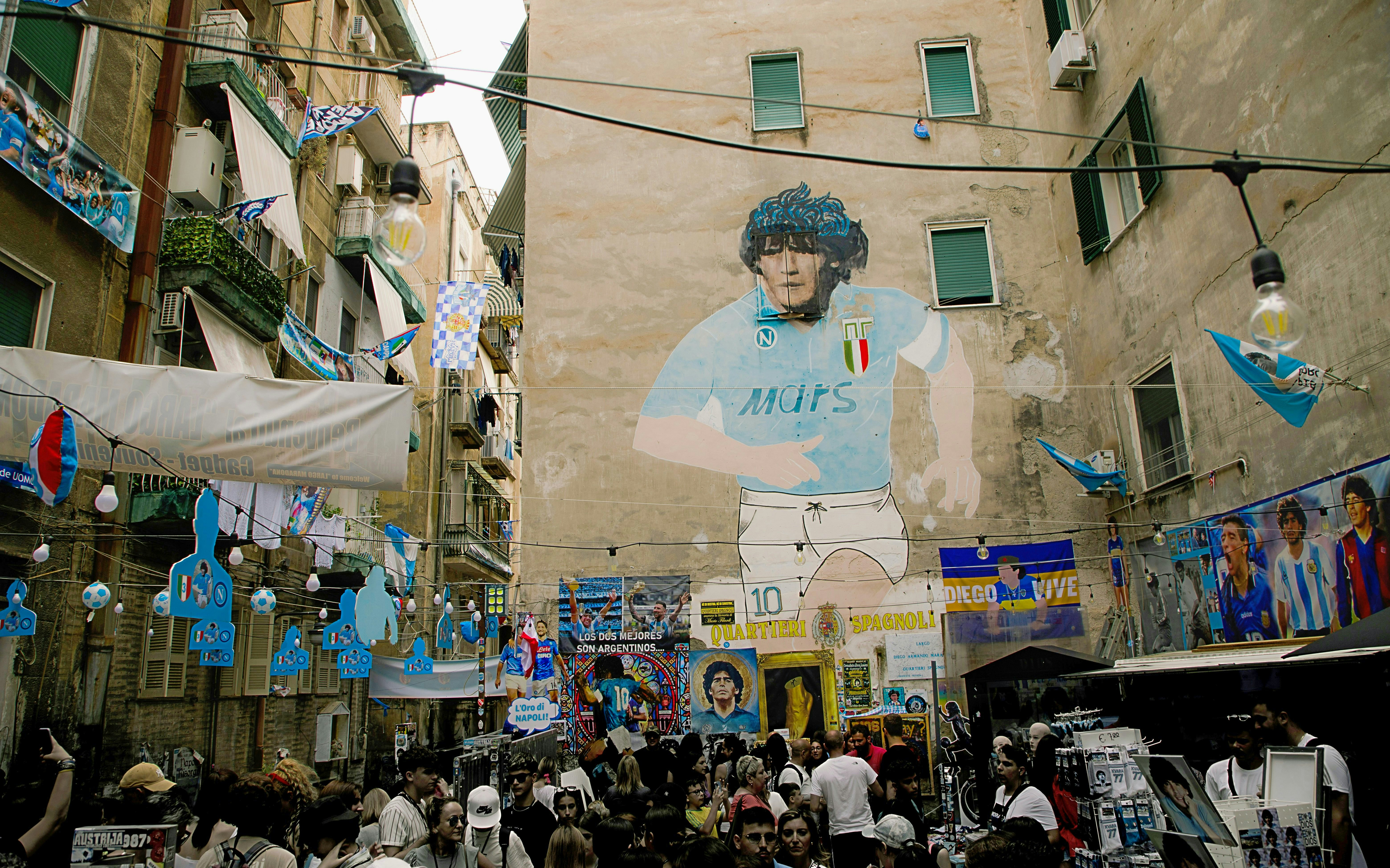 Mural of a famous footballer in Sanità District, Naples, with people exploring the vibrant street.