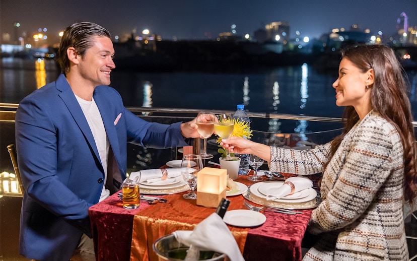 Couple toasting on Dubai Canal cruise at night with city lights in background.