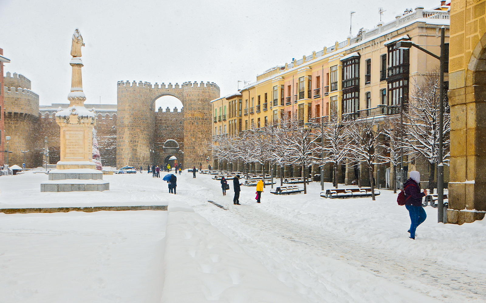Avila city square in winter, snow-covered ground, people walking, historic walls in background.