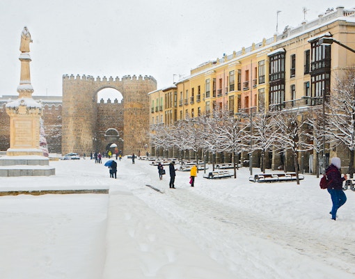Avila city square in winter, snow-covered ground, people walking, historic walls in background.