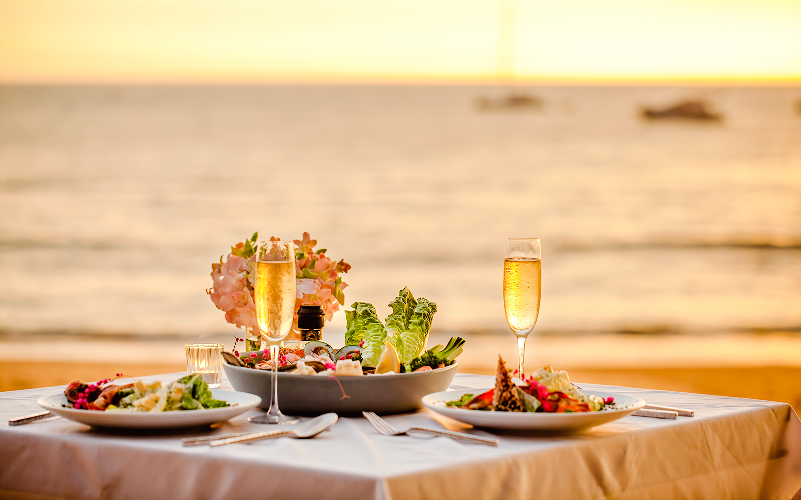 Romantic sunset dinner setup with champagne and salads by the ocean.
