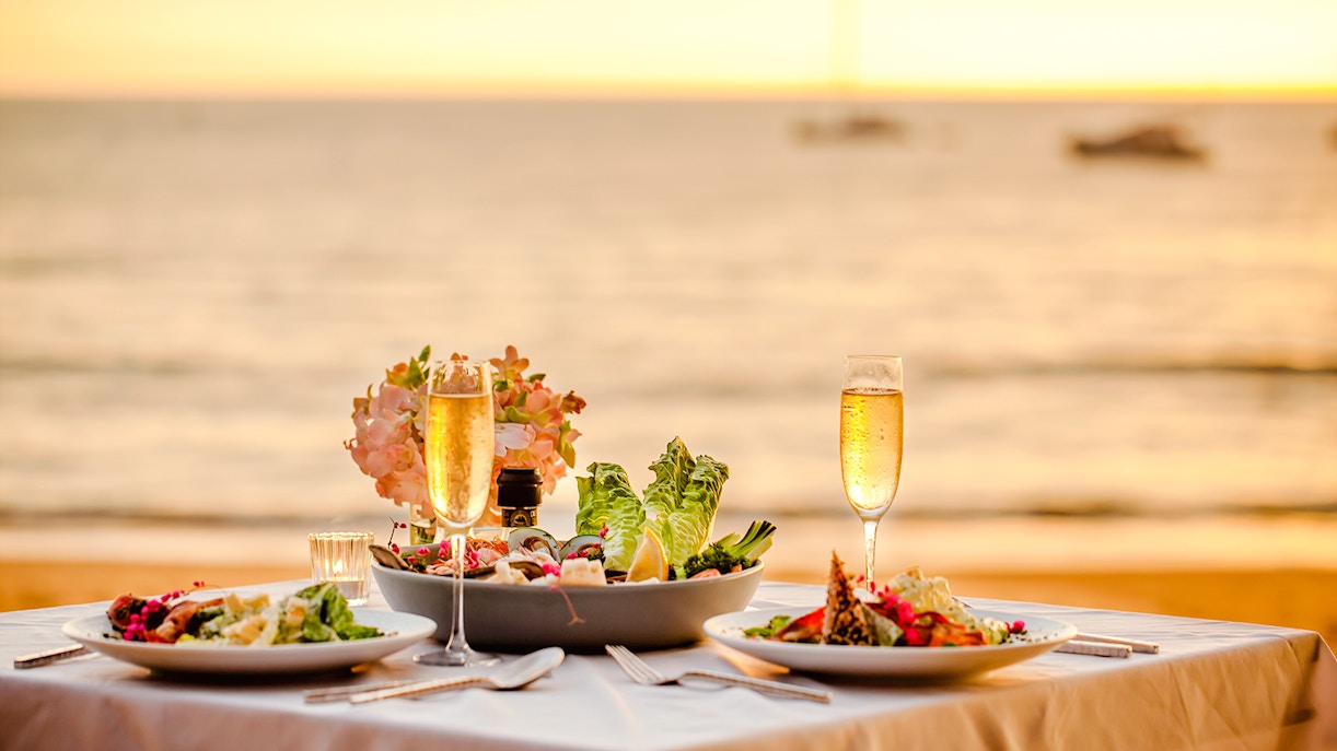 Romantic sunset dinner setup with champagne and salads by the ocean.