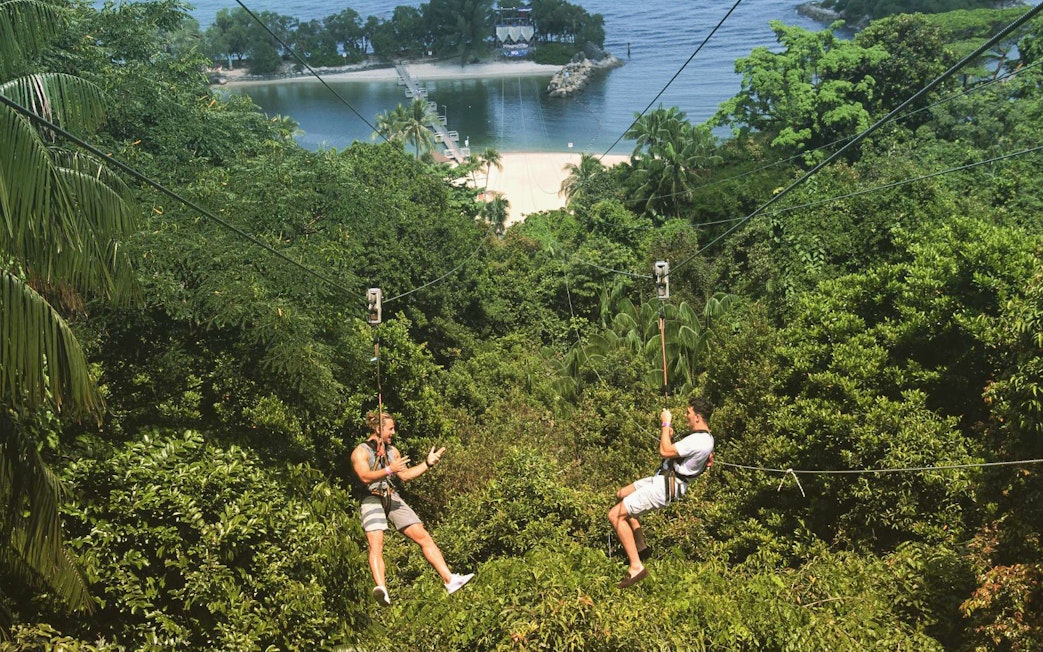 Two people ziplining over lush greenery at Mega Adventure, Sentosa, Singapore.
