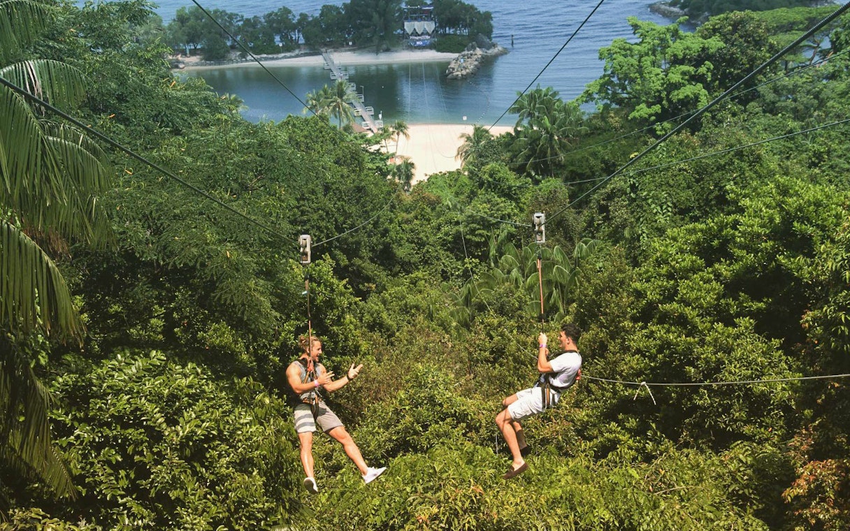 Two people ziplining over lush greenery at Mega Adventure, Sentosa, Singapore.