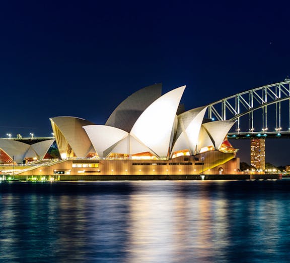 Sydney Opera House and Harbour Bridge illuminated at night, viewed from the water.