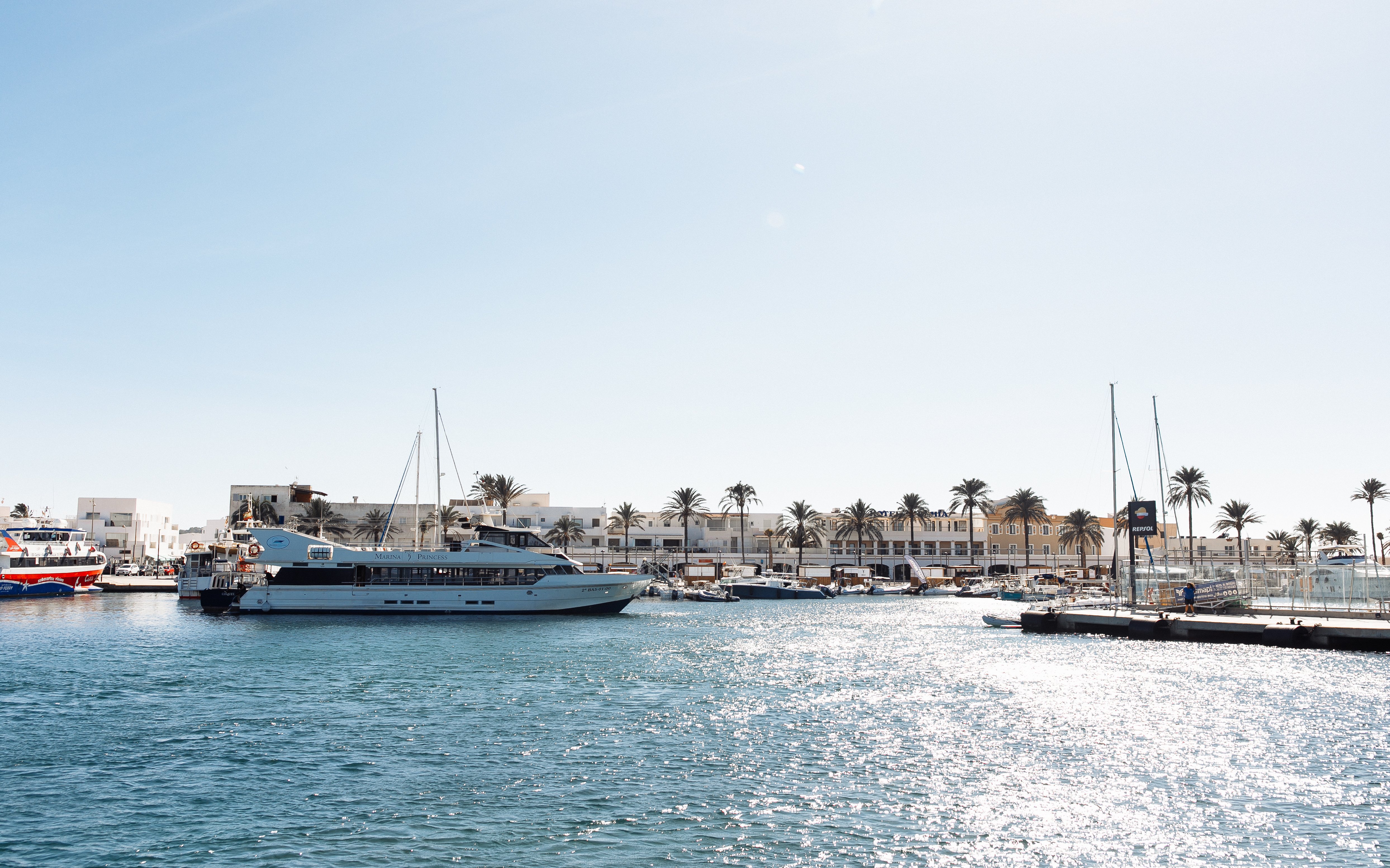 Formentera port view from a boat with docked yachts and palm-lined buildings.