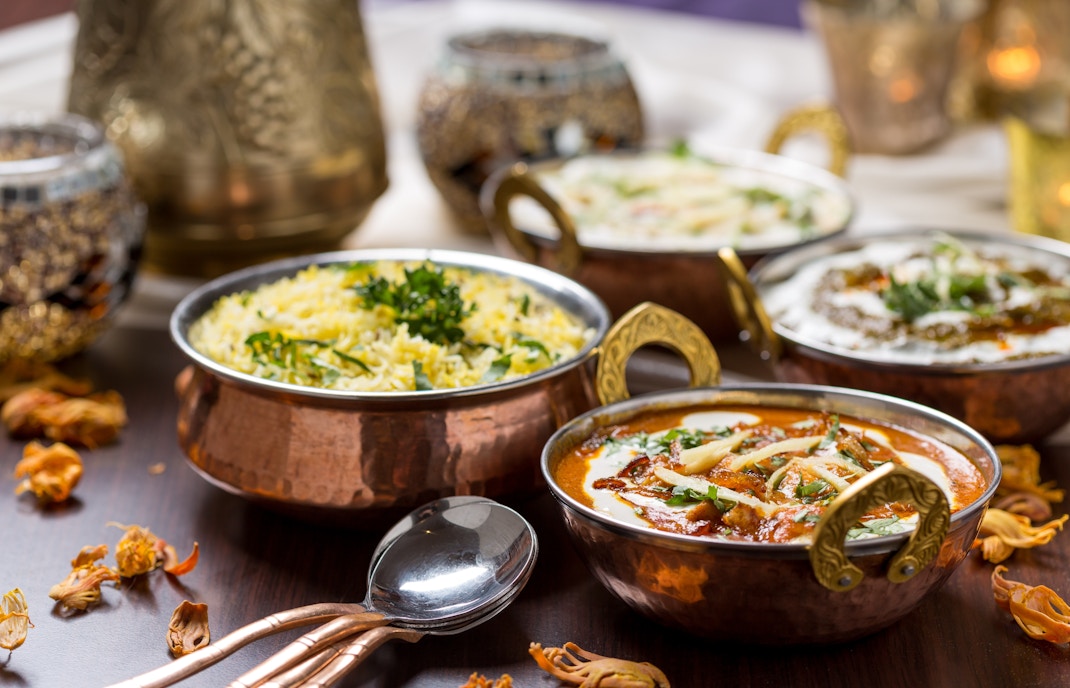 Copper bowls with Indian curry, rice, and yogurt dishes on a wooden table.