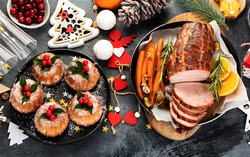 Sliced roast ham with carrots and oranges, surrounded by festive decorations and holiday pastries.