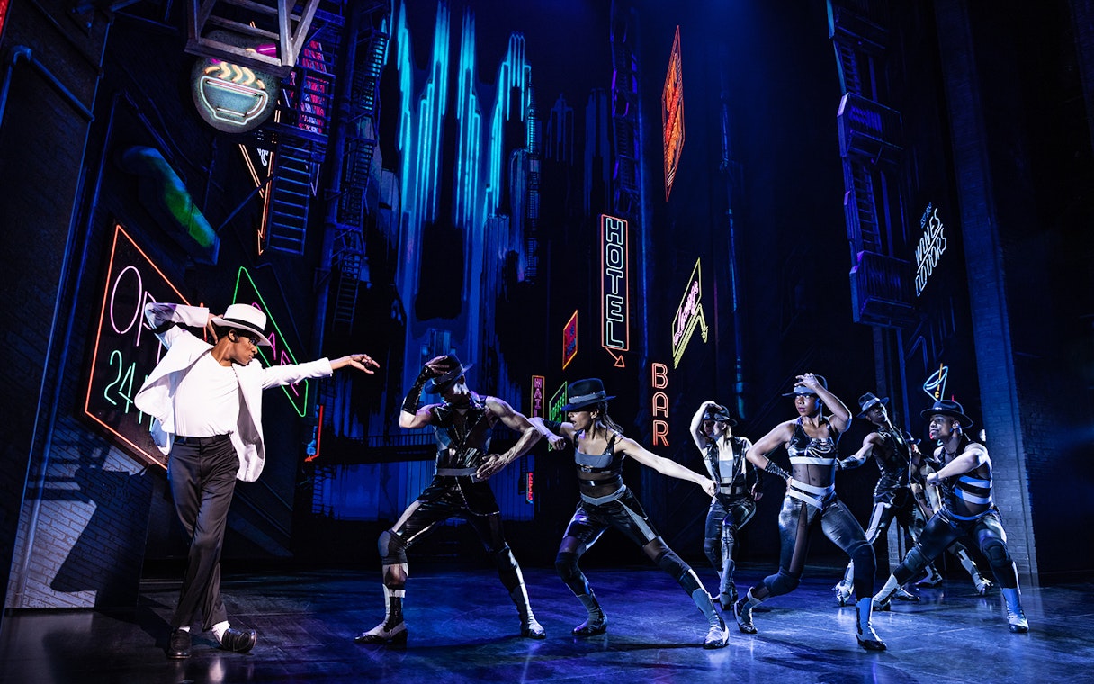 Performers dancing on stage at MJ Live tribute concert with neon city backdrop.