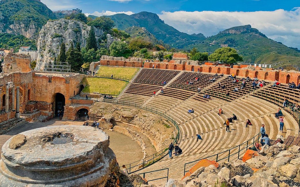 Taormina Ancient Theatre with visitors seated, surrounded by scenic hills.