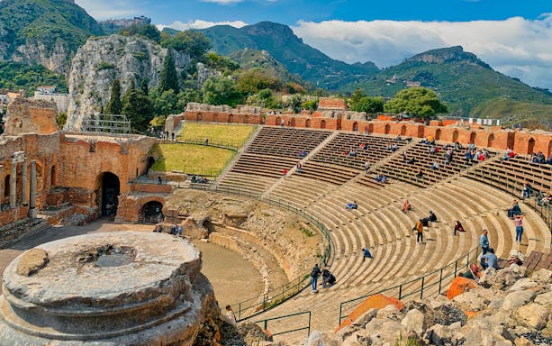 Taormina Ancient Theatre with visitors seated, surrounded by scenic hills.