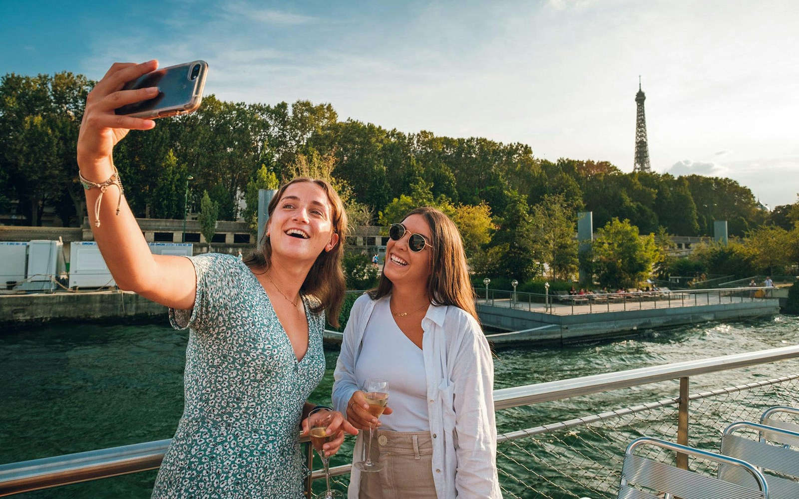 Young women taking selfies on a Seine River cruise with Vedettes de Paris, Eiffel Tower in background.