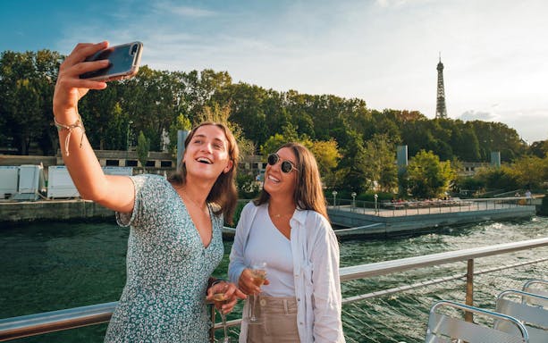 Young women taking selfies on a Paris cruise with Eiffel Tower in the background.