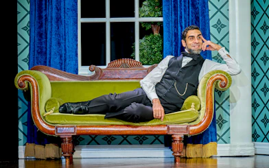 Man in period costume reclining on a vintage green sofa in a theatrical setting for "Oh Mary!