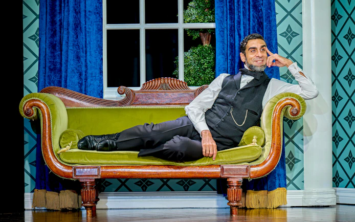 Man in period costume reclining on a vintage green sofa in a theatrical setting for "Oh Mary!