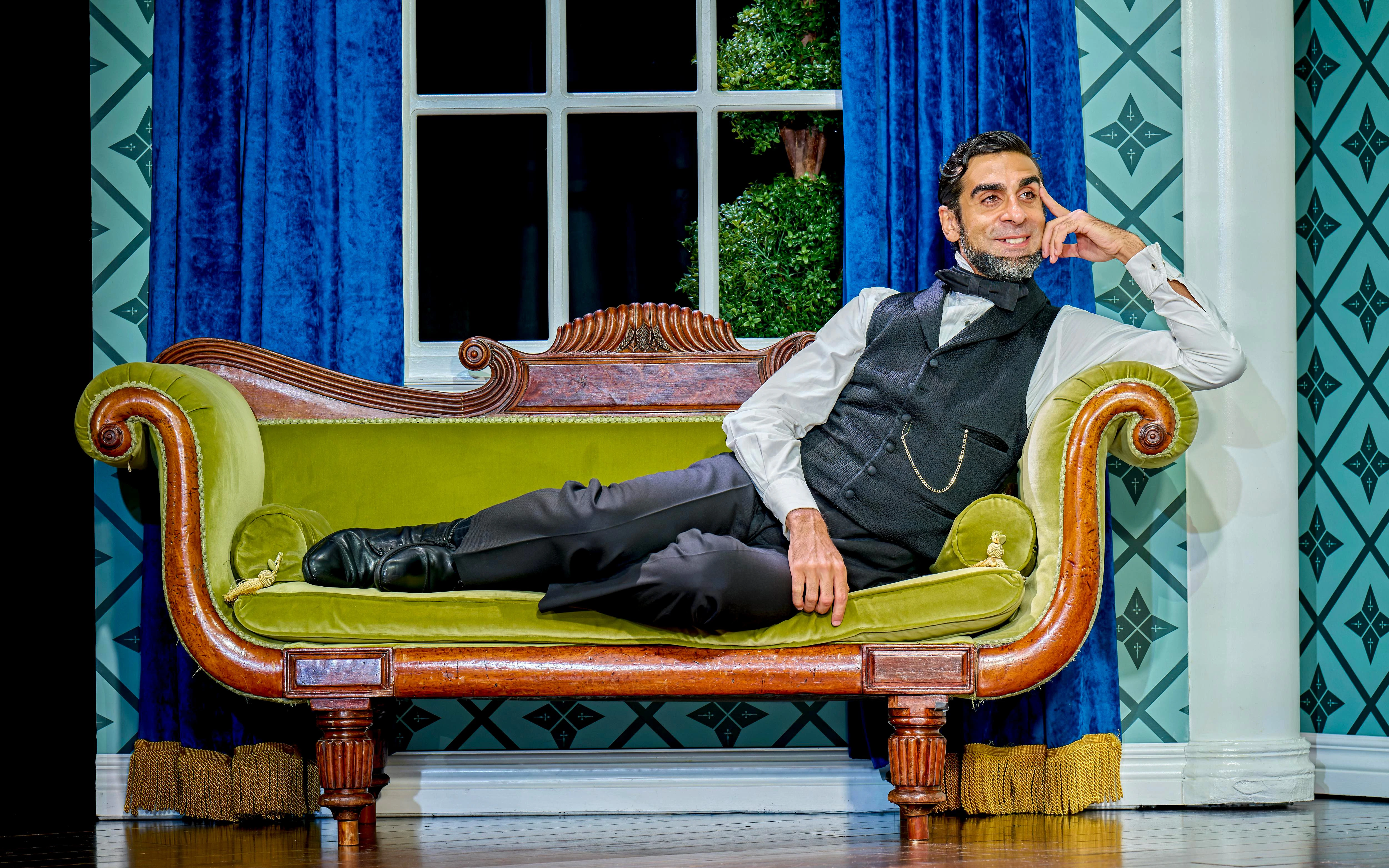 Man in period costume reclining on a vintage green sofa in a theatrical setting for "Oh Mary!